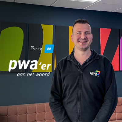 Perry, medewerker van PWA, staat lachend voor een kleurrijke wanddecoratie met de tekst ‘pwa’er aan het woord’.