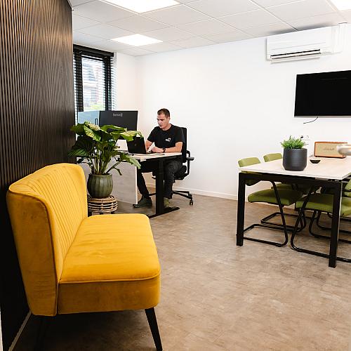 PWA-medewerker werkt achter een bureau in een modern en licht kantoor met een gele bank, groene stoelen en planten als interieuraccenten.