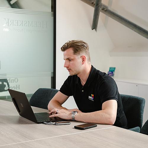 Man in PWA-bedrijfskleding werkt geconcentreerd op een laptop aan een vergadertafel in een modern kantoor met glazen wanden en industriële details.