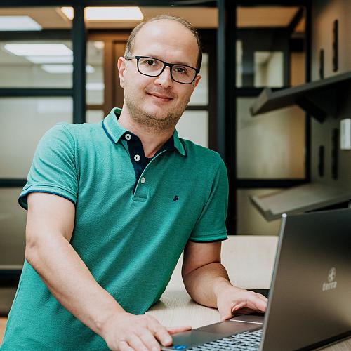 Man met bril werkt achter een laptop in een modern kantoor met glazen wanden en warme verlichting.
