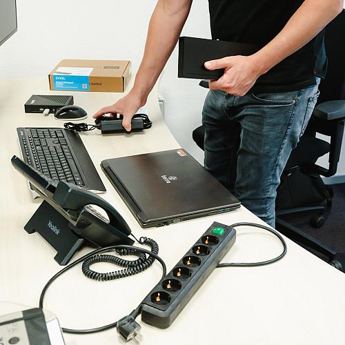 Man installeert apparatuur op een bureau met een gesloten laptop, toetsenbord, telefoontoestel en stekkerdoos in een kantooromgeving.