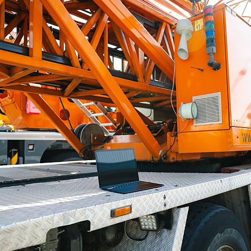 Laptop staat op het werkplatform van een grote oranje hijskraan, klaar voor gebruik bij werkzaamheden op locatie.