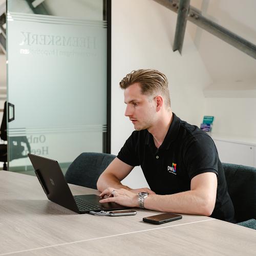 Man in PWA-bedrijfskleding werkt geconcentreerd op een laptop aan een vergadertafel in een modern kantoor met glazen wanden en industriële details.