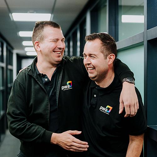  Twee PWA-collega’s lachen samen in de kantoorhal, met een arm om elkaars schouder, als teken van teamspirit en fijne werksfeer.