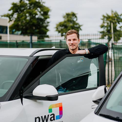 Man in PWA-bedrijfskleding leunt tegen de open deur van een witte PWA-auto op een bedrijventerrein, met bomen en hekwerk op de achtergrond.