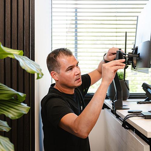  PWA-medewerker stelt een monitor af op een verstelbare arm in een modern kantoor met veel daglicht en groene planten.