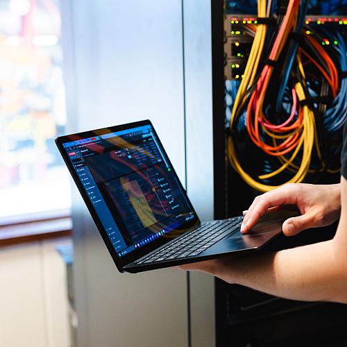 Persoon werkt met een laptop voor een open patchkast vol kleurrijke netwerkkabels en knipperende lampjes.