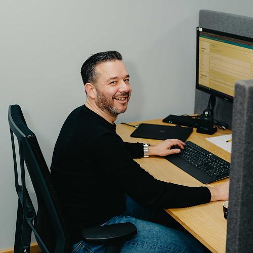 Lachende man zit achter een bureau en werkt op een computer in een moderne kantooromgeving met grijze schermwanden.