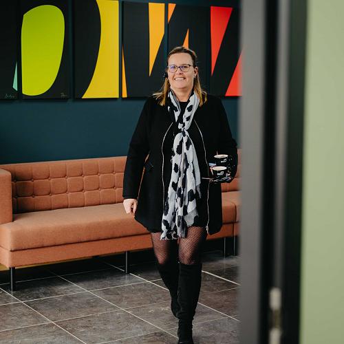 Vrouw die een kantoorruimte binnenloopt met een koffie in de hand, voor een moderne wand met kleurrijke kunst en een roze bank.