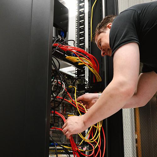 Man werkt geconcentreerd aan een serverkast en sluit kleurrijke netwerkkabels aan in een technische ruimte.