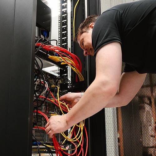 Man werkt geconcentreerd aan een serverkast en sluit kleurrijke netwerkkabels aan in een technische ruimte.