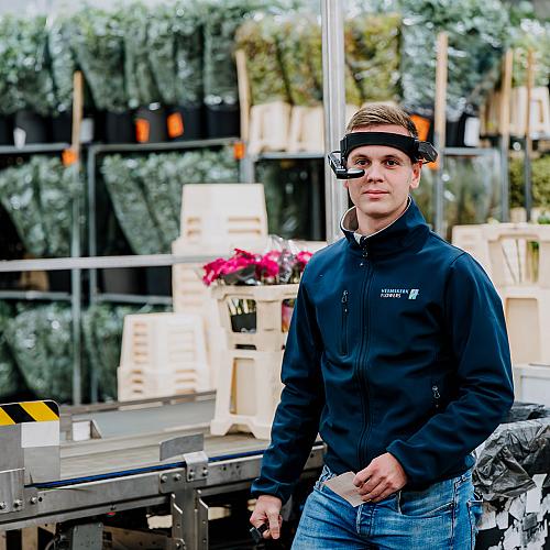 Medewerker met een slimme headset loopt door een hal vol bloemenkarren en bereidt orders voor binnen de productieomgeving.