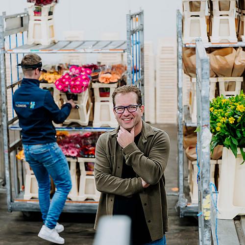 Man die lachend poseert in een bloemenhal, terwijl op de achtergrond een medewerker bloemen in karren sorteert voor verwerking en distributie.