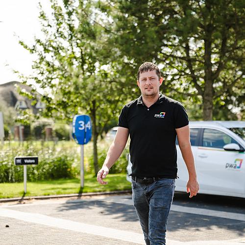 PWA-medewerker loopt buiten naast een PWA-auto op een zonnige dag.