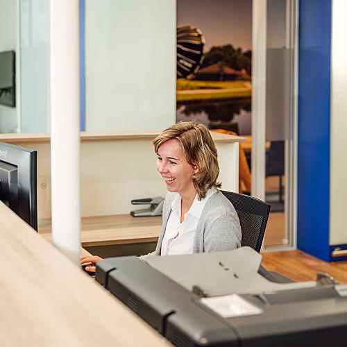 Receptioniste achter de balie werkt lachend aan haar computer in een modern kantoor.
