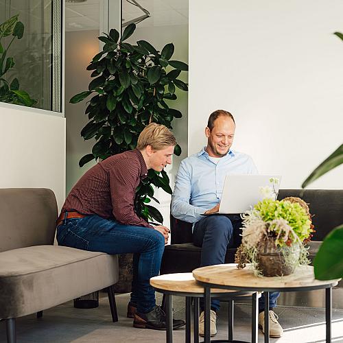 Twee collega’s zitten in een loungehoek en bespreken iets op een laptop tussen groene planten.