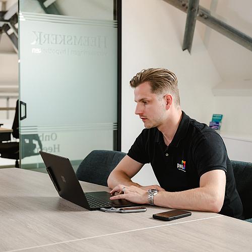 PWA-medewerker die geconcentreerd werkt op een laptop aan een vergadertafel, met smartphone en notitieboekje binnen handbereik.