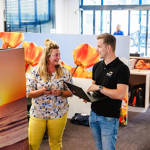 Collega’s overleggen staand in een kantoorruimte met laptop en decoratieve wandpanelen.