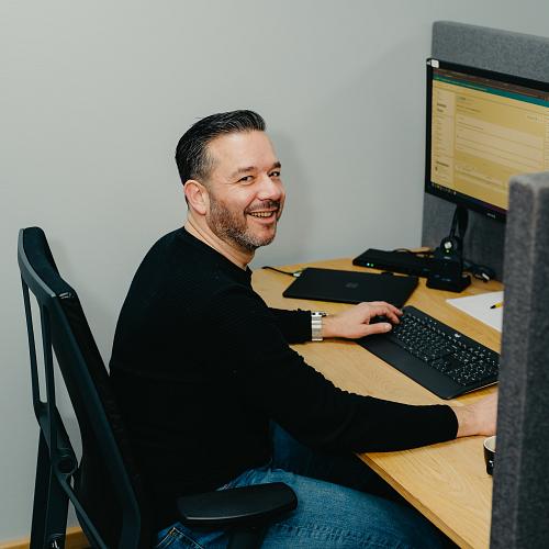 PWA-medewerker werkt aan zijn computer op kantoor en kijkt glimlachend richting de camera.