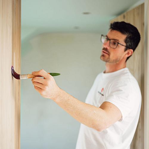 Schilder die met een kwast een houten wand afwerkt tijdens renovatiewerkzaamheden in een binnenruimte.