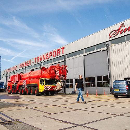 Bedrijfshal van Verschoor Kranen Transport met grote rode mobiele kraan op het buitenterrein en medewerker die langs het pand loopt.