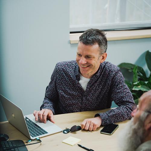 PWA-medewerker bespreekt IT-oplossing met klant aan tafel met laptop en smartphone op kantoor.