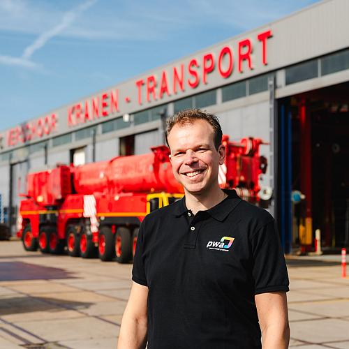 Lachende man in PWA-shirt staat voor een werkplaats van een kraan- en transportbedrijf met opvallende rode en blauwe voertuigen op de achtergrond.