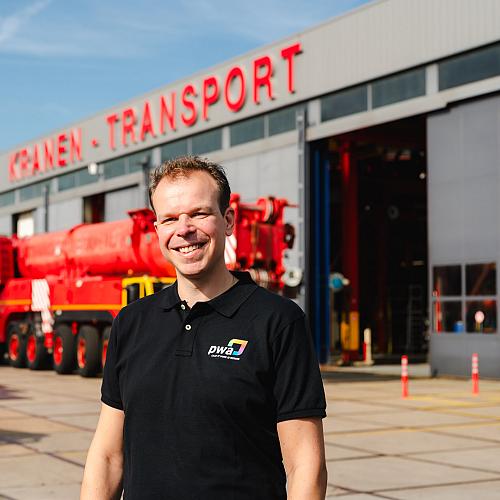 Lachende man in PWA-shirt staat voor een werkplaats van een kraan- en transportbedrijf met opvallende rode en blauwe voertuigen op de achtergrond.