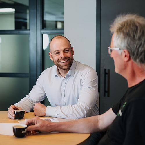 Twee mannen zitten aan een tafel met koffiekopjes in een moderne kantoorruimte. De man in een lichtblauw overhemd glimlacht naar zijn gesprekspartner, die op de voorgrond te zien is.