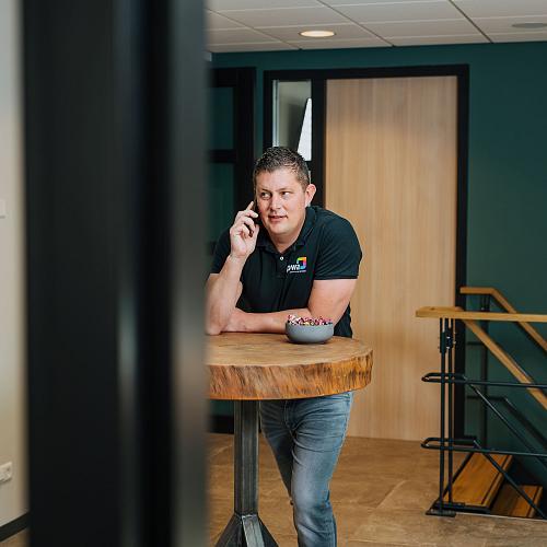 Man voert een telefoongesprek terwijl hij leunt op een hoge houten tafel in een moderne kantoorruimte.
