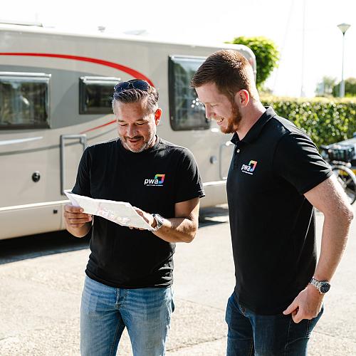 Twee mannen in PWA-kleding bekijken samen een plattegrond of document op straat, met een camper en auto op de achtergrond.