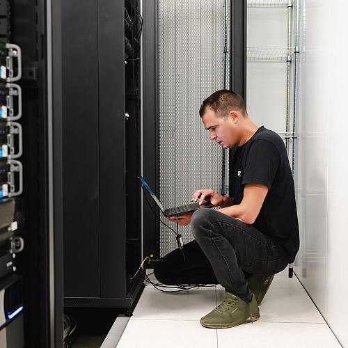 IT-specialist werkt gehurkt met een laptop voor een serverkast in een datacenter, gefocust bezig met netwerk- of systeembeheer.