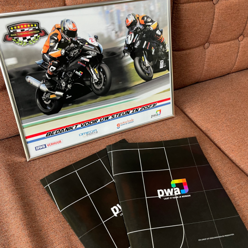 Foto van een bank met twee zwarte PWA-mappen en een ingelijste actiefoto van motorracers met diverse sponsoren, waaronder PWA.