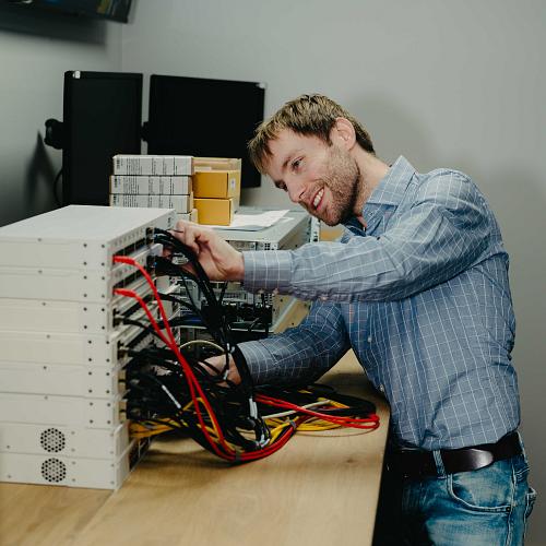 Man werkt met een glimlach aan een stapel netwerkapparatuur op een houten werktafel, terwijl hij kabels aansluit in een technische ruimte.