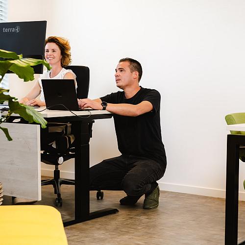 Vrouw zit lachend achter een bureau terwijl een man gehurkt naast haar aan een laptop werkt, in een lichte kantoorruimte met groene stoelen en veel planten.