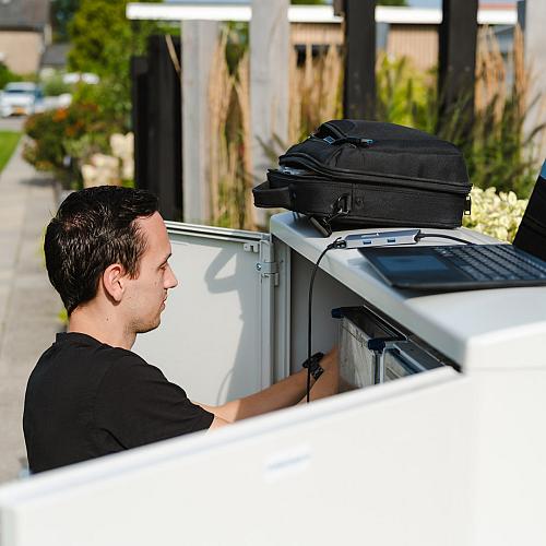 Monteur werkt aan een buitenkast in een woonwijk, met een open laptop en rugtas erop geplaatst voor registratie of diagnose.