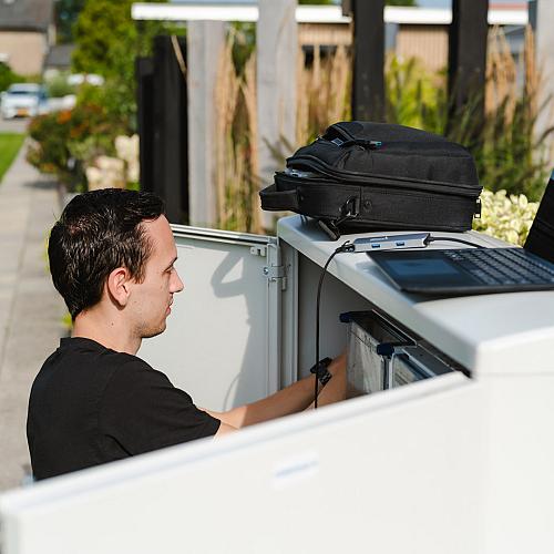 Monteur werkt aan een buitenkast in een woonwijk, met een open laptop en rugtas erop geplaatst voor registratie of diagnose.