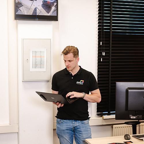 Man in PWA-bedrijfskleding werkt staand op een laptop in een kantoorruimte met beeldschermen en gesloten jaloezieën.