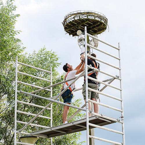 Twee monteurs in veiligheidsharnas werken op een steiger aan apparatuur onder een hoog platform in een groene buitenomgeving.
