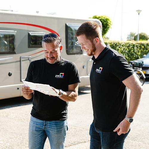 Twee mannen in PWA-kleding bekijken samen een plattegrond op straat, met een camper en auto op de achtergrond op een zonnige dag.
