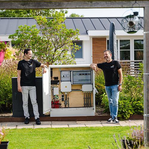 Twee mannen in PWA-shirt staan glimlachend aan weerszijden van een open technische kast in een groene tuin bij een modern gebouw.