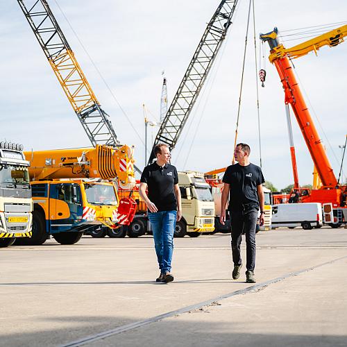 Twee mannen in PWA-kleding lopen over een kraanterrein met grote hijskranen en vrachtwagens op de achtergrond.