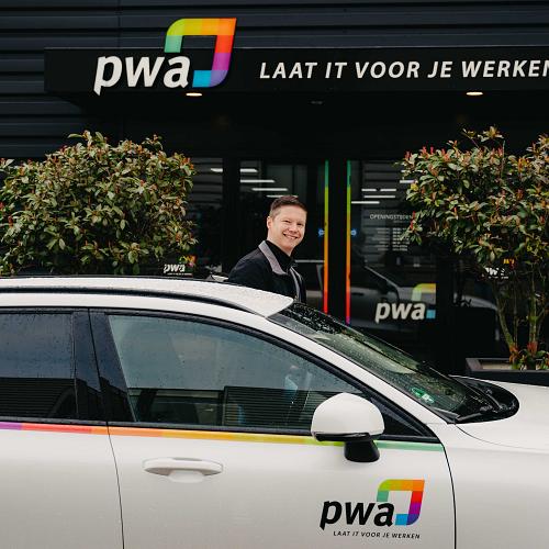 Man stapt lachend uit een auto met PWA-logo voor het hoofdkantoor van PWA met de slogan ‘Laat IT voor je werken’ boven de ingang.