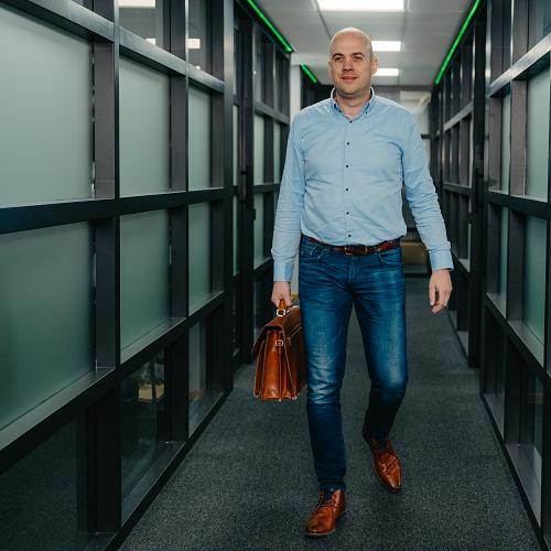 Man loopt door een modern kantoorpand met glazen wanden, gekleed in een lichtblauw overhemd en spijkerbroek, en draagt een leren aktetas.