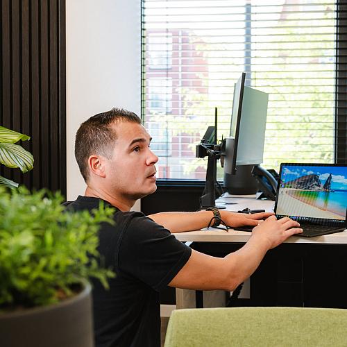 Man in zwart shirt werkt op een laptop aan een bureau in een modern kantoor met veel licht en groene planten.