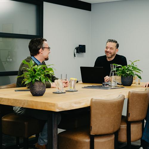 3 lachende manne om vergadertafel met groene plantjes en een glas water.