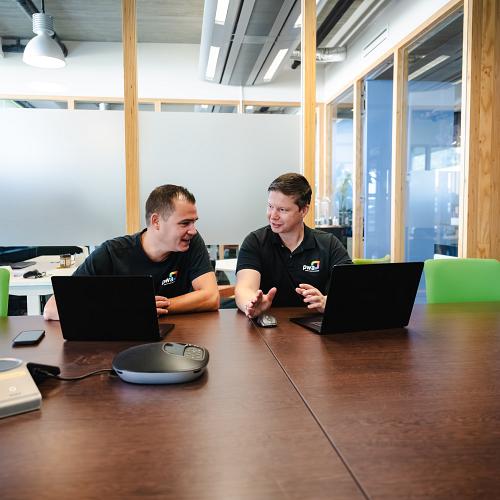 Twee lachende mannen achter een vergadertafel met groene stoelen, met twee laptops voor hun in gesprek.