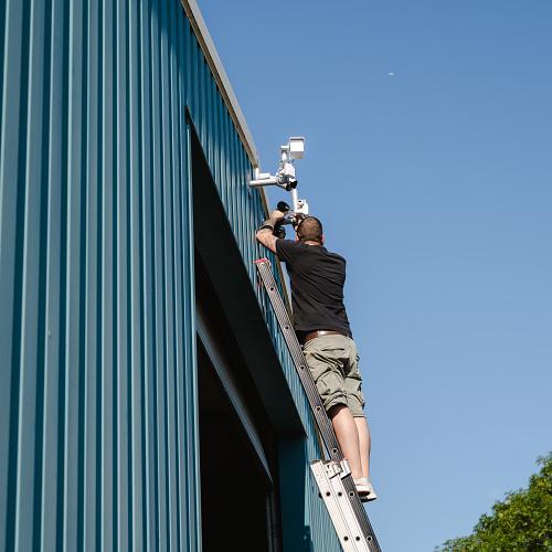 Man in zwart shirt staand op een ladder voor een blauw gebouw, die bezig is met een beveiligingscamera ophangen.