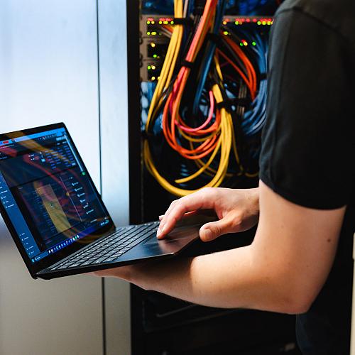 Persoon werkt met een laptop voor een open patchkast vol kleurrijke netwerkkabels en knipperende lampjes.