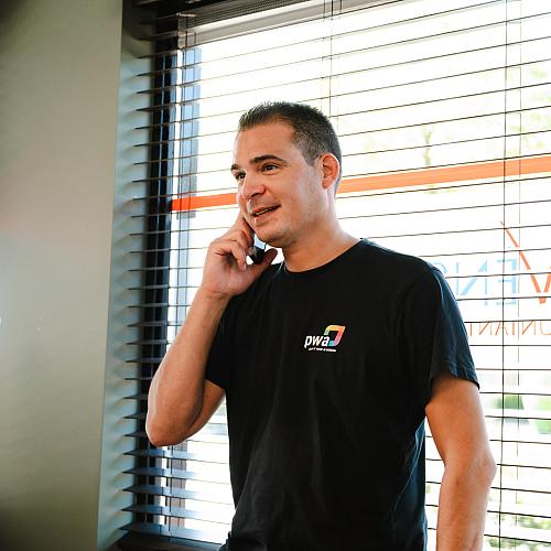 Man in PWA-shirt belt glimlachend bij het raam van een kantoor met jaloezieën en het logo van Novinco op de ruit.